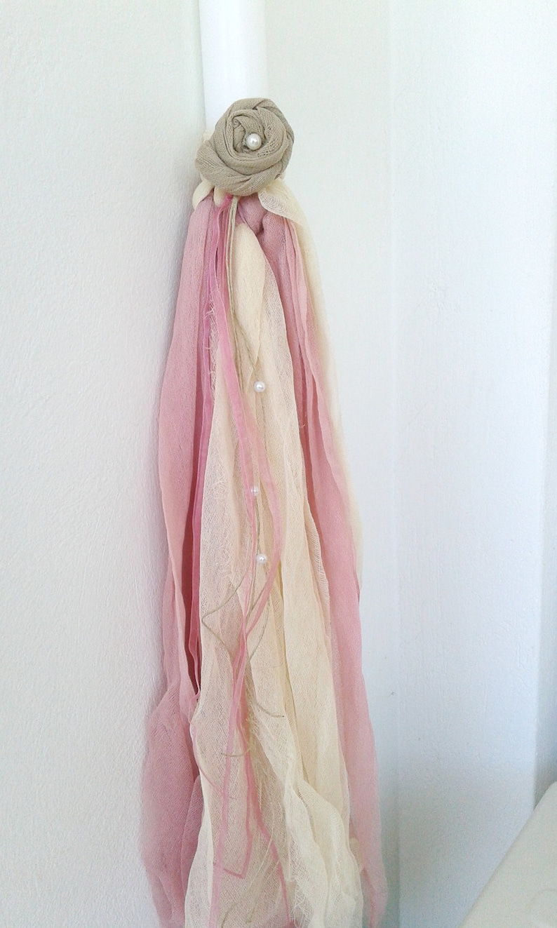 May include: A white candle with a pink and cream fabric tassel and a beige fabric flower with a pearl accent.