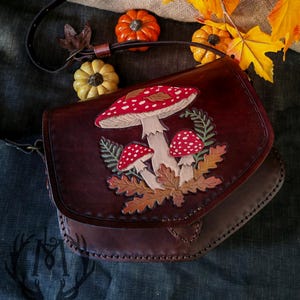 Pode incluir: Bolsa transversal de couro castanho escuro com aba, decorada com um motivo de cogumelos vermelhos, brancos e bege. A bolsa tem uma alça longa e ajustável. Folhas de outono e pequenas abóboras são dispostas ao redor.