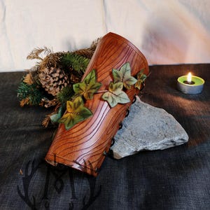 May include: A brown leather arm cuff with a wood grain design and green ivy leaves. The cuff is decorated with small metal rivets.