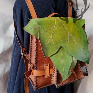 Op de afbeelding: Een bruine leren rugzak met een groot groen blad dat op de voorkant is genaaid. De rugzak heeft een klep met een gespsluiting en een klein vakje aan de zijkant. De banden zijn verstelbaar en gemaakt van bruin leer.