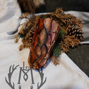 Puede incluir: Un brazalete de cuero hecho a mano con un diseño texturizado único en tonos marrón y burdeos, asegurado con pequeños remaches dorados. El brazalete se exhibe sobre una superficie blanca con un logotipo de cuernos gris y rodeado de agujas de pino.