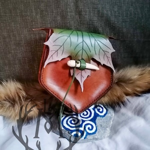 Puede incluir: Una bolsa de cuero marrón con un diseño de hoja verde y un cierre de hueso. La bolsa está colgada de un cordón blanco.