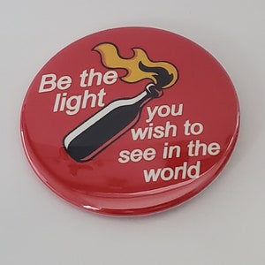 May include: A red button with the text "Be the light you wish to see in the world." A black bottle with a flame is in the center. The button is round and has a glossy finish.