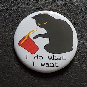 May include: A white button with a black cat illustration tipping over a red cup, with yellow liquid spilling out. The text "I do what I want" is printed below the image. The button is round and has a black border.