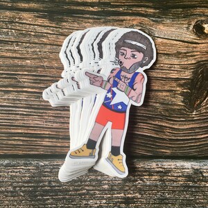 May include: A stack of stickers featuring a cartoon character with an afro, wearing a blue and white jersey with a star, red shorts, and yellow sneakers. The character is pointing with one hand and holding a microphone with the other. The sticker has a white background.