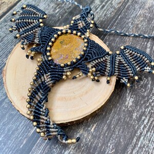 May include: A black and brown macrame dragon pendant with a gold-toned gemstone in the center. The pendant is hanging from a brown cord and is displayed on a wooden surface.