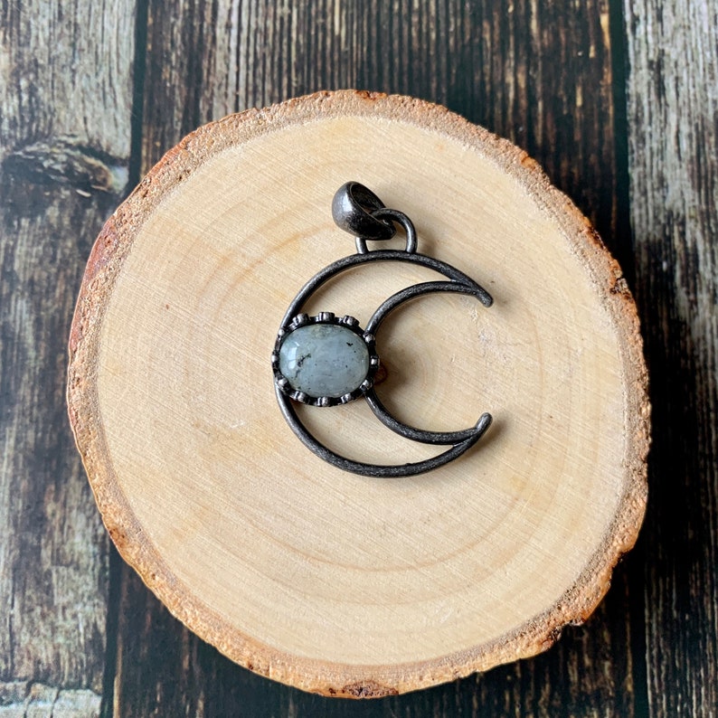 K&ouml;nnte beinhalten: Ein silberner Halbmond-Anh&auml;nger mit einem grauen Mondstein-Cabochon in der Mitte. Der Anh&auml;nger liegt auf einer Holzoberfl&auml;che.
