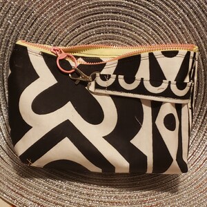 May include: A rectangular black and white patterned cosmetic bag with a pink zipper and a matching pink clasp. The bag has a small wrist strap with a decorative design. The interior is yellow.