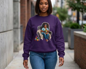 May include: A purple sweatshirt featuring an illustration of a person with an afro sitting in a chair and holding yarn. The illustration includes a blanket, yarn balls, and the person is wearing a blue shirt and jeans.
