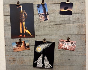 May include: A wooden board with various postcards attached with clips. The postcards feature a variety of images, including a bird, a beach scene, a comet, and a piano keyboard.