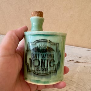May include: A green ceramic bottle with a cork stopper. The bottle has a label that reads "Bat Wing Tonic Elixir".