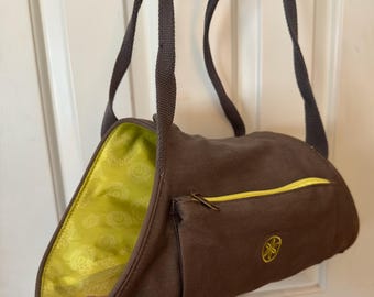 Pode incluir: Bolsa de ombro de lona marrom com zíper amarelo e um pequeno bolso frontal. A bolsa tem uma alça cinza e um interior verde brilhante com um padrão floral. Um logotipo amarelo é bordado na frente.