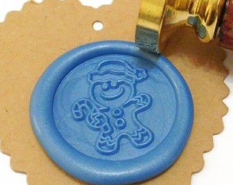 May include: A blue wax seal with a gingerbread man design is pressed onto a heart-shaped kraft paper tag. The seal is being applied with a gold-colored metal stamp with a wooden handle. The gingerbread man is wearing a hat and holding a candy cane.