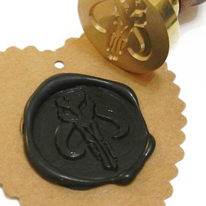 May include: A brass wax seal stamp with a wooden handle, next to a black wax seal impression on a brown paper tag. The seal features a stylized design. The stamp is angled towards the impression.