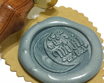 Puede incluir: Un sello de cera gris con el texto "Fly you to the moon" y un diseño de luna creciente. El sello está en una etiqueta de papel marrón y un mango de madera con una parte superior de latón.