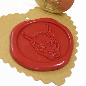 Könnte beinhalten: Ein goldfarbener Wachssiegel-Stempel mit einem detaillierten Design einer japanischen Dämonenmaske. Der Stempel wird verwendet, um ein rotes Wachssiegel mit dem gleichen Design zu erstellen.