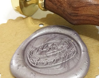 May include: A silver wax seal with an embossed design sits on a scalloped-edge kraft paper tag. A wooden handle with a brass head is positioned above the seal, suggesting its use for creating impressions.