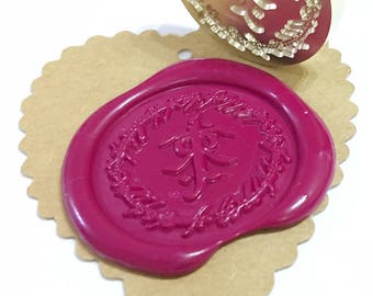 Puede incluir: Un sello de cera dorado con un diseño floral y caracteres chinos. El sello se utiliza para crear un sello de cera rojo con el mismo diseño y caracteres.