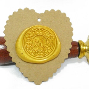 Pode incluir: Um selo de cera dourada com a letra "K" no centro, rodeado por um design ornamentado. O selo está em uma etiqueta de papel kraft em forma de coração. A alça de madeira e o carimbo de latão são visíveis.