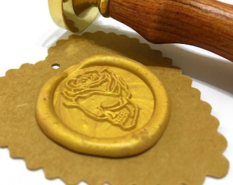 Puede incluir: Un sello de cera dorado con un diseño de rosa en una etiqueta marrón en forma de corazón. El sello de cera está hecho de cera dorada y tiene un diseño en relieve de una rosa.