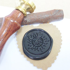 May include: A wax seal stamp with a wooden handle and a brass head. The black wax seal features a sun and pentagram design, resting on a heart-shaped piece of brown paper. A wooden stick is also present.