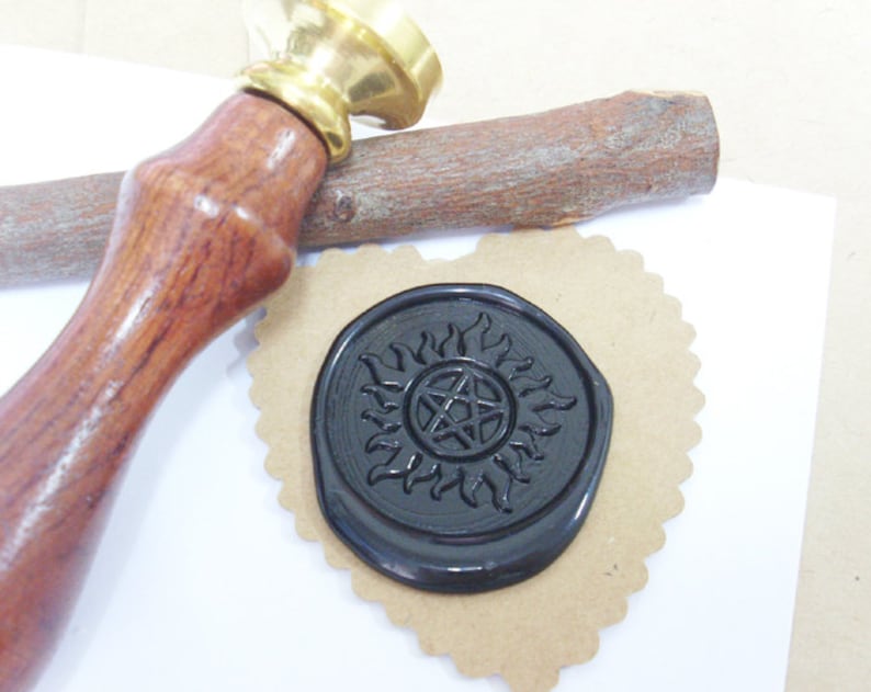 May include: A wax seal stamp with a wooden handle and a brass head. The black wax seal features a sun and pentagram design, resting on a heart-shaped piece of brown paper. A wooden stick is also present.
