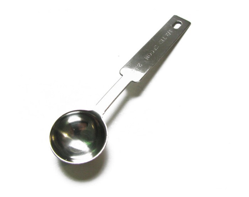 Puede incluir: Una cuchara medidora de plata con un cuenco redondeado y un mango largo. El mango tiene el texto "1/2 TSP" impreso en &eacute;l.