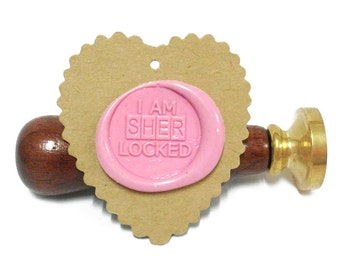May include: A pink wax seal with the text "I AM SHER LOCKED" on a heart-shaped tag attached to a wooden and brass wax seal stamp.