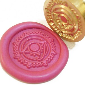 May include: A close-up of a red wax seal with a decorative eye design. The seal is made with a brass stamp with a similar design.