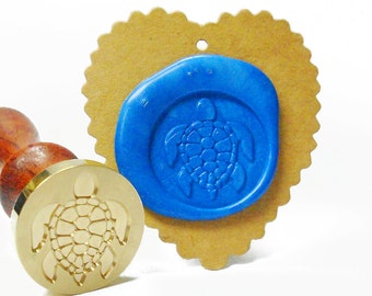 May include: A brass wax seal stamp with a turtle design next to a blue wax seal impression on a heart-shaped tag. The stamp has a wooden handle. The blue wax seal is on a brown tag with a scalloped edge.