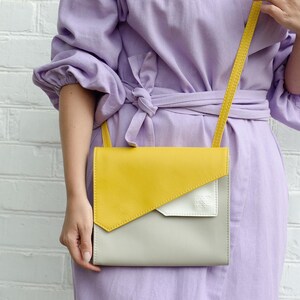 May include: A yellow, gray, and white leather crossbody bag with a long strap. The bag is worn by a person wearing a lavender dress.