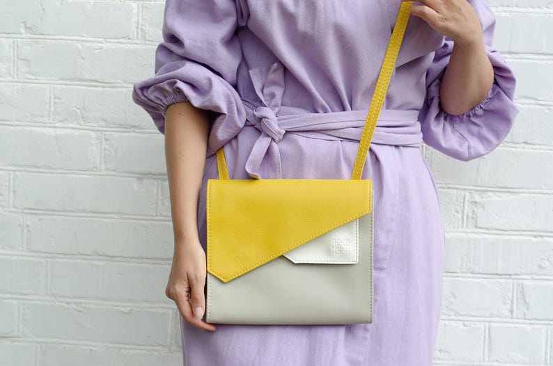 May include: A yellow, gray, and white leather crossbody bag with a long strap. The bag is worn by a person wearing a lavender dress.
