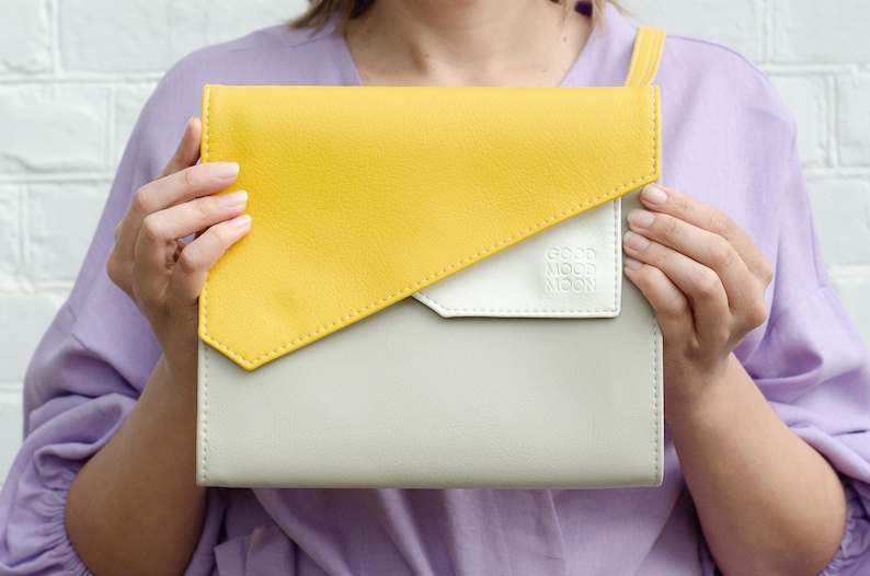 May include: A yellow and white leather clutch with a flap closure. The clutch has a white patch with the text "GOOD MOOD NOW" on it.