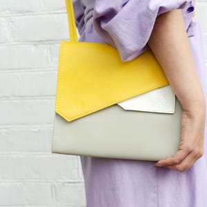 May include: A yellow and gray leather shoulder bag with a white flap. The bag is being held by a person wearing a lavender shirt.