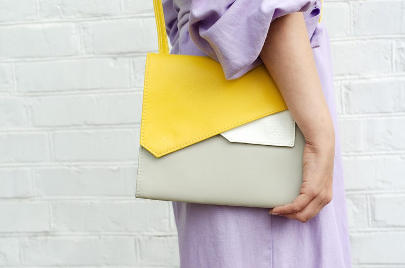 May include: A yellow and gray leather shoulder bag with a white flap. The bag is being held by a person wearing a lavender shirt.