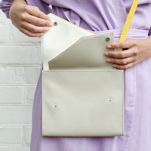 May include: A light gray leather crossbody bag with a yellow strap. The bag is open and the inside is white.