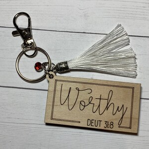 May include: Wooden keychain with a white tassel, a silver ring, and a red jewel. The keychain has the word "Worthy" engraved on it, along with the scripture reference "DEUT 31:8".