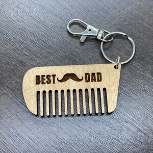 May include: Wooden comb keychain with the text "BEST DAD" and a mustache graphic. The comb has a silver key ring attached.