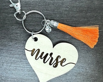 May include: A wooden heart-shaped keychain with the word "nurse" engraved on it. The keychain has a silver ring and a silver lobster clasp. A small orange tassel hangs from the ring.