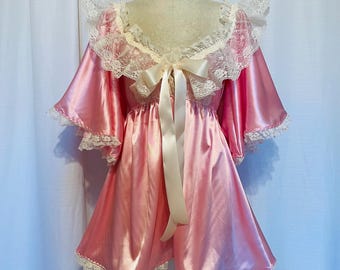 May include: A pink satin robe with a cream-colored lace trim and a matching bow. The robe has short sleeves and a ruffled collar. The garment is displayed on a mannequin against a white backdrop.