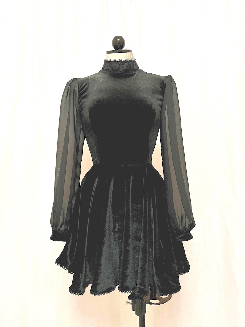 May include: A black velvet dress with sheer long sleeves and a high neck. The dress has a fitted bodice and a flared skirt with a scalloped hem.