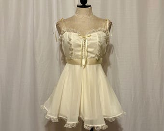 May include: A cream-colored dress on a mannequin. The dress features a bodice with lace trim and a ribbon tie, thin straps, and a flared skirt. A yellow ribbon accents the waist. The hemline is trimmed with lace.