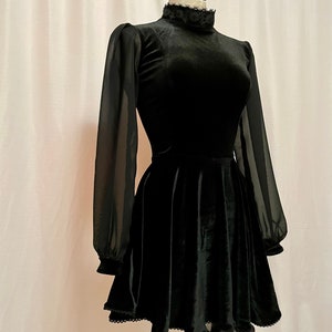 May include: A black velvet dress with sheer long sleeves and a high neck. The dress has a fitted bodice and a flared skirt with a scalloped hem.