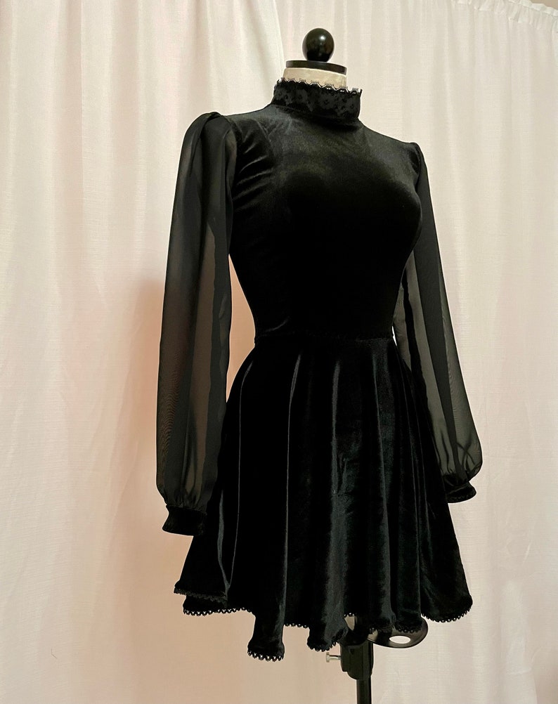 May include: A black velvet dress with sheer long sleeves and a high neck. The dress has a fitted bodice and a flared skirt with a scalloped hem.