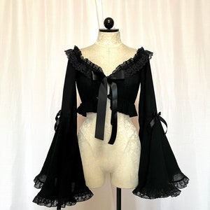 May include: A black cropped top with long, bell sleeves and lace trim. The top features a lace neckline and a black ribbon tie at the front.
