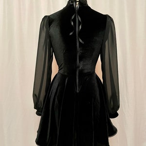 May include: Black velvet dress with sheer long sleeves and a high neckline. The dress has a fitted bodice and a flared skirt with a scalloped hem.