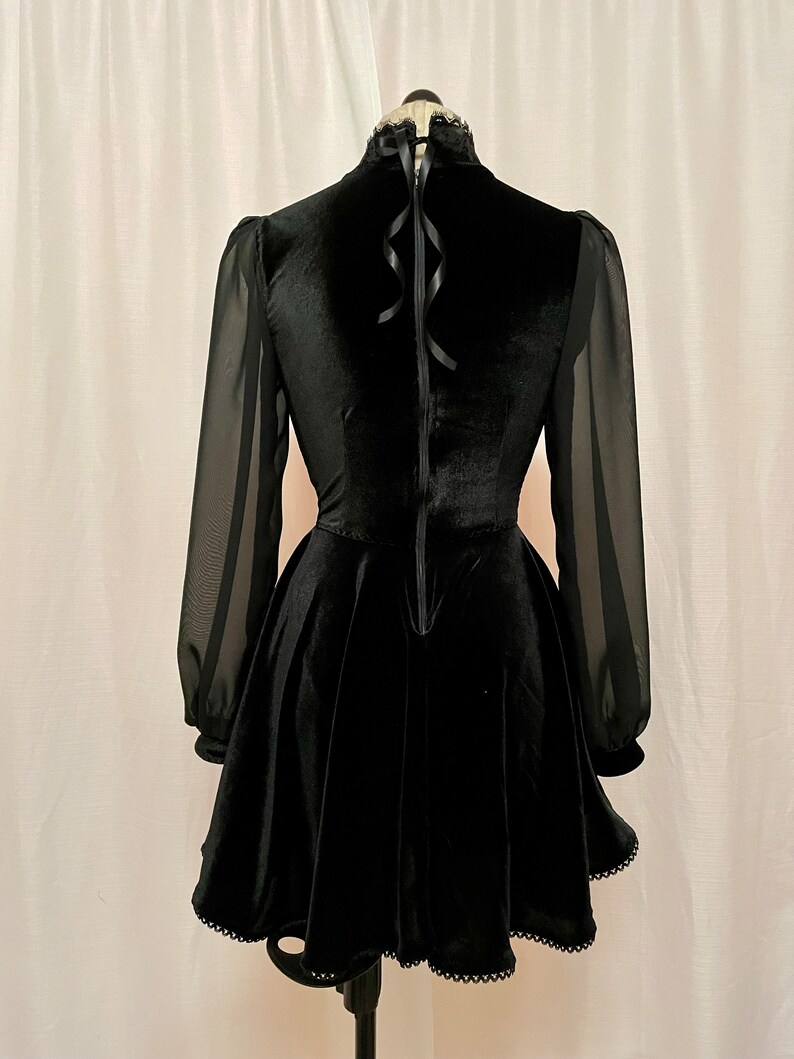 May include: Black velvet dress with sheer long sleeves and a high neckline. The dress has a fitted bodice and a flared skirt with a scalloped hem.