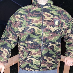 Puede incluir: Chaqueta de forro polar de camuflaje con cremallera completa, con un patrón de verde, marrón y negro. La chaqueta tiene una cremallera negra y un parche en el pecho con el texto "D.M.D.C." y un gráfico.