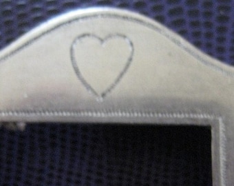 May include: A silver-toned picture frame with a heart-shaped design at the top. The frame has a rectangular opening for a photo.