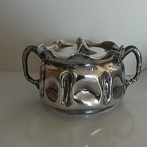 May include: A silver sugar bowl with a decorative, scalloped lid and two handles. The bowl has a textured, ornate design.
